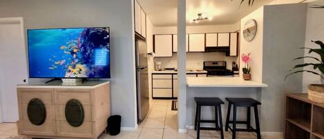 A modern living room with a flat-screen TV, a wooden cabinet, and a kitchen area with a bar stool.