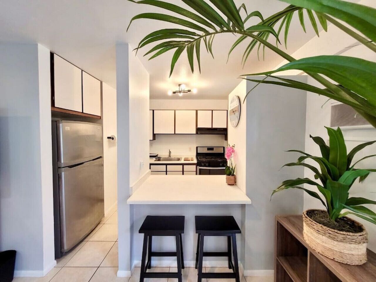 A modern kitchen with a stainless steel refrigerator, white cabinets, a central island with two black stools, and potted plants.
