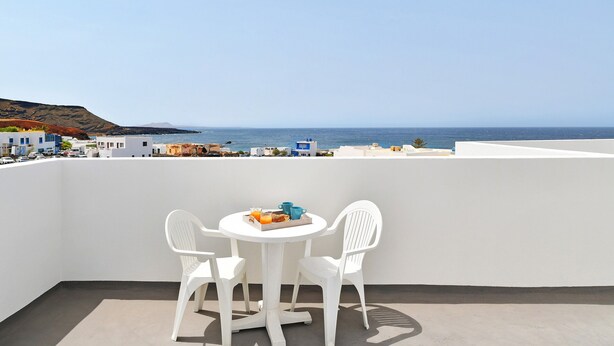 Un balcon avec une table et des chaises blanches, un plateau avec de la nourriture et des boissons, et une vue sur la mer et les bâtiments.