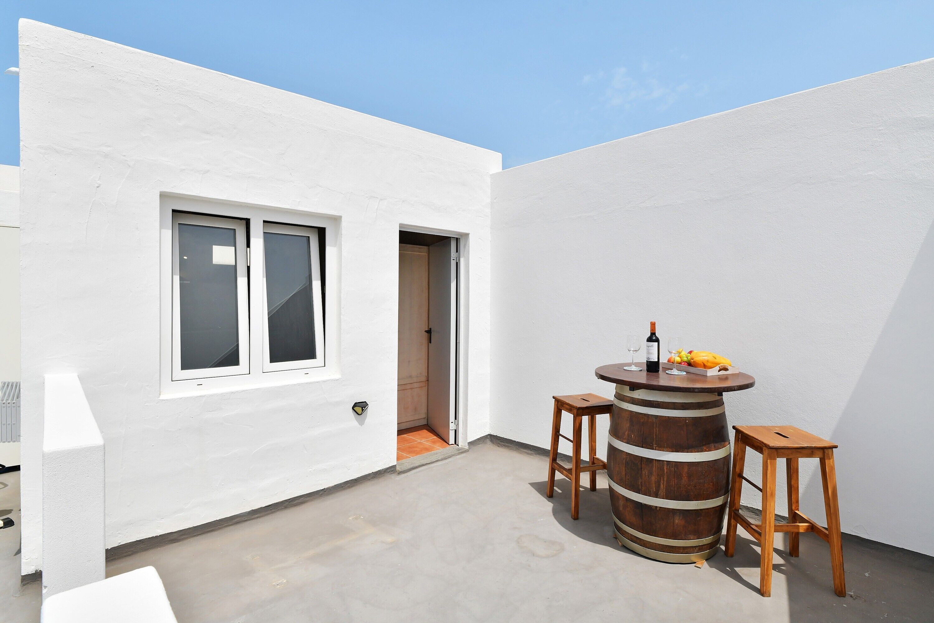 A white exterior wall with a window and a door, a concrete floor, and a wooden barrel stool with a bottle and fruit on top.