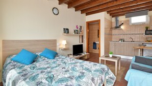 Une chambre comprenant un lit, un bureau, une chaise, une télévision, une petite table avec une plante, et un coin cuisine avec un four à micro-ondes et des placards.