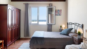 Un dormitorio con cama, armario, ventana con cortinas, mesita de noche con lámpara y un cuadro en la pared.