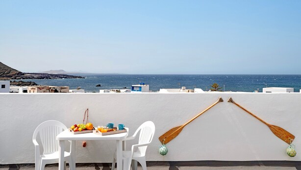 Una mesa con frutas y bebidas, dos sillas blancas y dos remos de madera en un balcón con vista al mar.