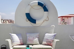 Um sofá de vime branco com almofadas estampadas, um círculo de boia e vista para edifícios ao fundo.