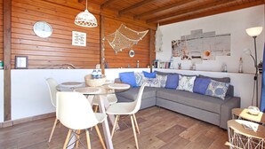 Un interior acogedor con techo de madera, una mesa redonda de vidrio con sillas, un sofá gris con cojines estampados y una pared decorada con motivos náuticos.