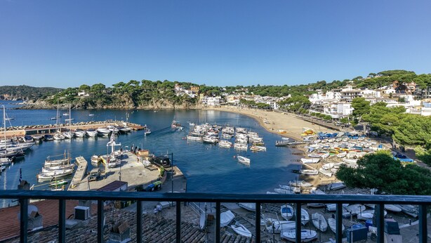 Una marina costera con barcos amarrados, una playa de arena y edificios al fondo.