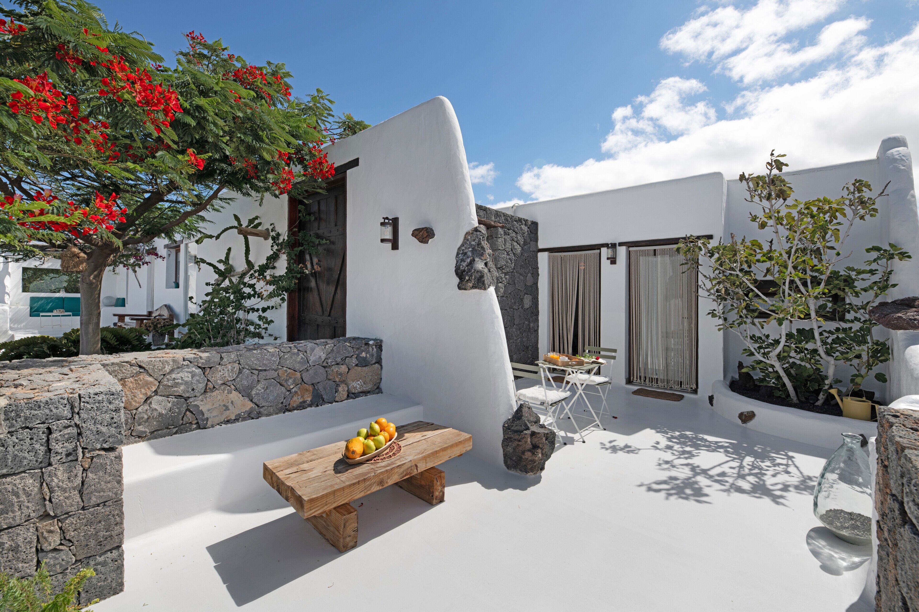 A white-walled outdoor area with a stone wall, a wooden bench with fruit, a small table, and a potted plant.
