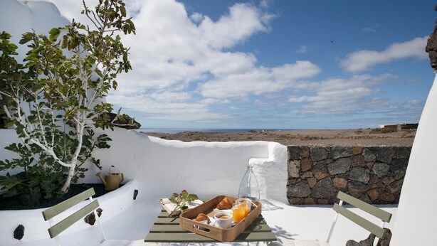 Habitación con paredes blancas, una pared de piedra, un banco, una bandeja con comida y bebida, y vista al mar.