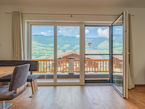 A spacious room with a wooden dining table and chairs, large sliding glass doors leading to a balcony with a wooden railing, and a view of a mountainous landscape. - Group accommodation in panoramic location (Mittersill)