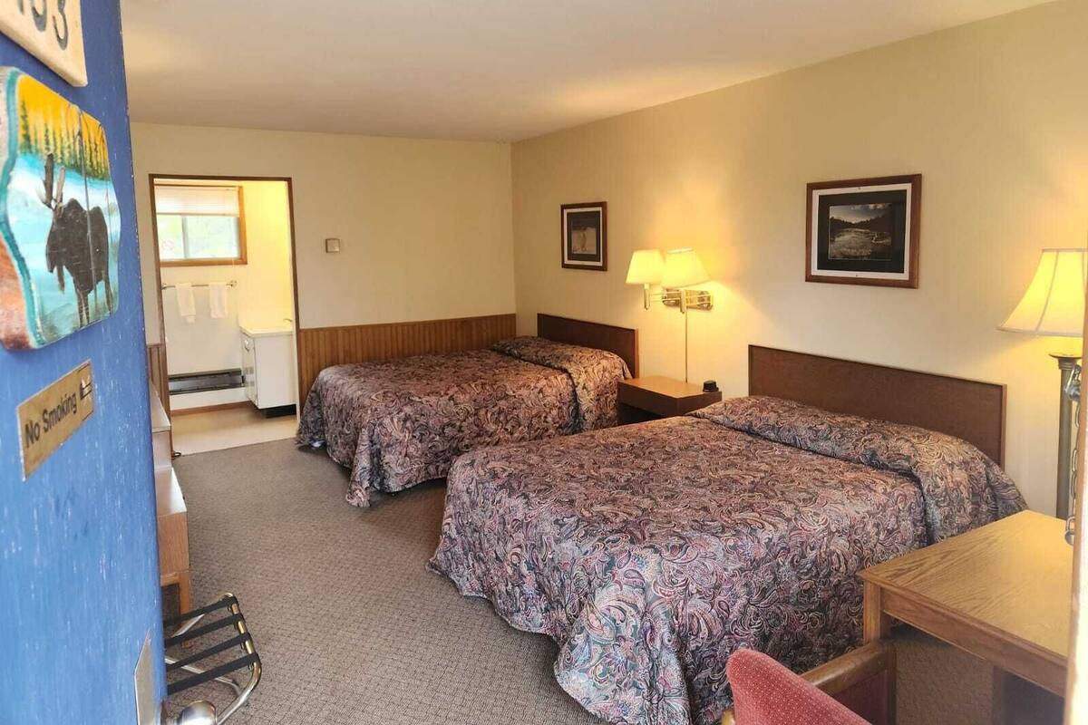 A hotel room with two beds, a desk, a chair, a lamp, and a painting on the wall.