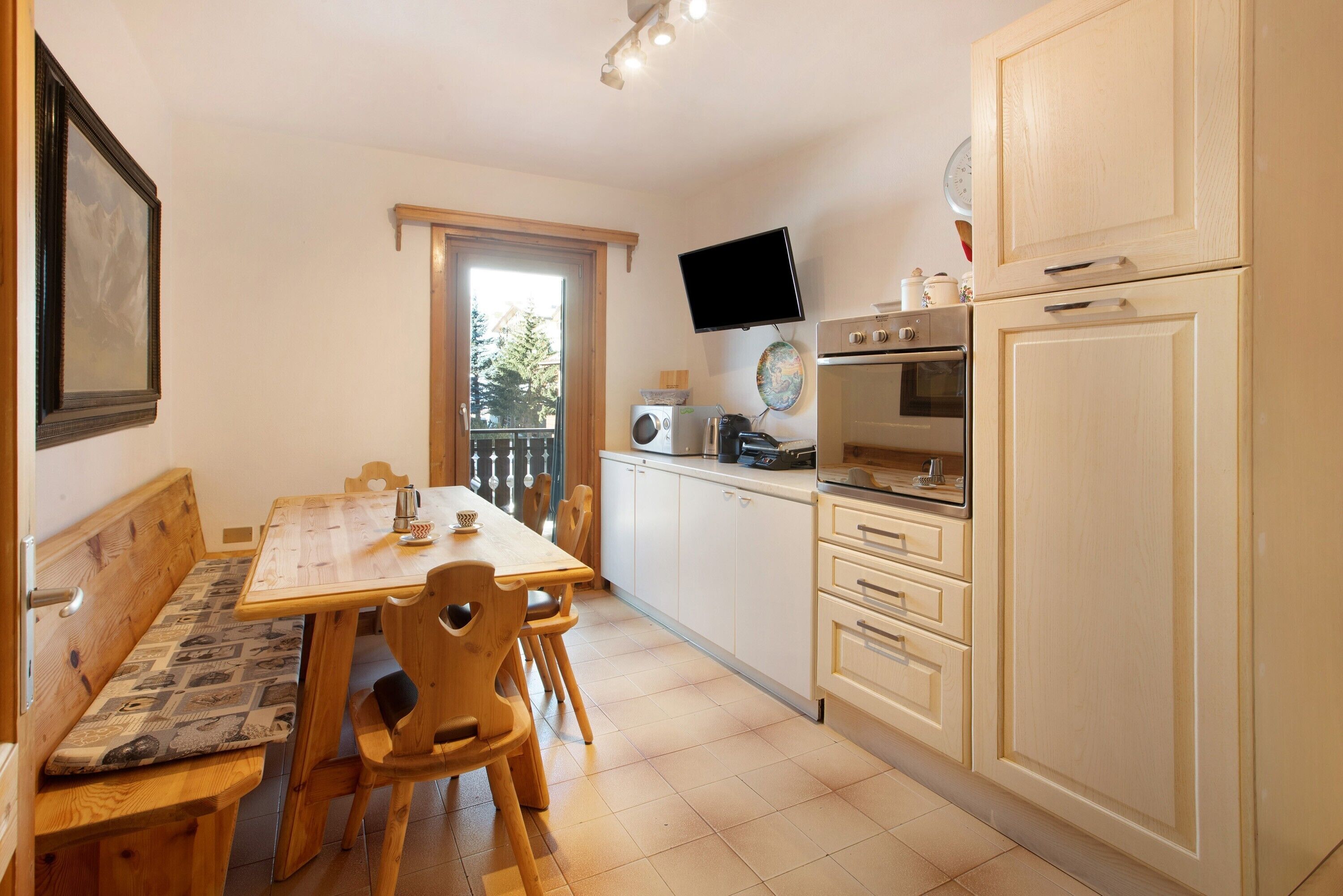 A kitchen with a dining table, chairs, a bench, cabinets, a television, and a window.