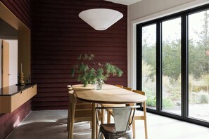 A dining area with a wooden table and chairs, a white pendant light, and a view of the outdoors through large windows.
