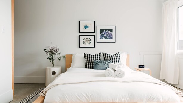 A modern bedroom with a wooden bed, white bedding, a small bedside table with a lamp, and two framed pictures on the wall. - Starbase - Waterfront Home w Pickleball Court (Prince Edward)