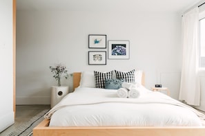 Un dormitorio moderno con una cama de madera, ropa de cama blanca, una mesita de noche con lámpara y dos cuadros en la pared.