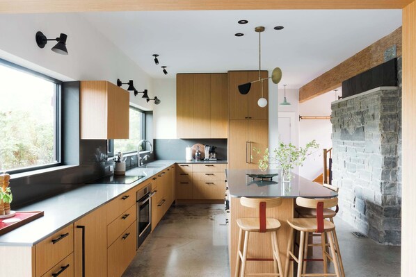 A modern kitchen with wooden cabinets, a central island with stools, a stone fireplace, and pendant lighting.