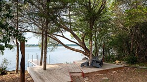A wooden deck with a bench and a table, overlooking a calm body of water. - Starbase - Waterfront Home w Pickleball Court (Prince Edward)