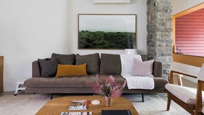A living room with a brown sofa, a coffee table with magazines, a vase with pink flowers, and a framed landscape picture on the wall. - Starbase - Waterfront Home w Pickleball Court (Prince Edward)