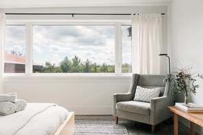 A modern hotel room with a large window, a grey armchair, a wooden side table, and a bed with white bedding.