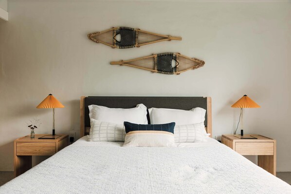 Una cama bien hecha con un edredón blanco texturizado, dos mesitas de noche de madera con lámparas y dos elementos decorativos en la pared.