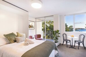 A hotel room with a bed, a small table with chairs, a balcony with a view, and a large window.
