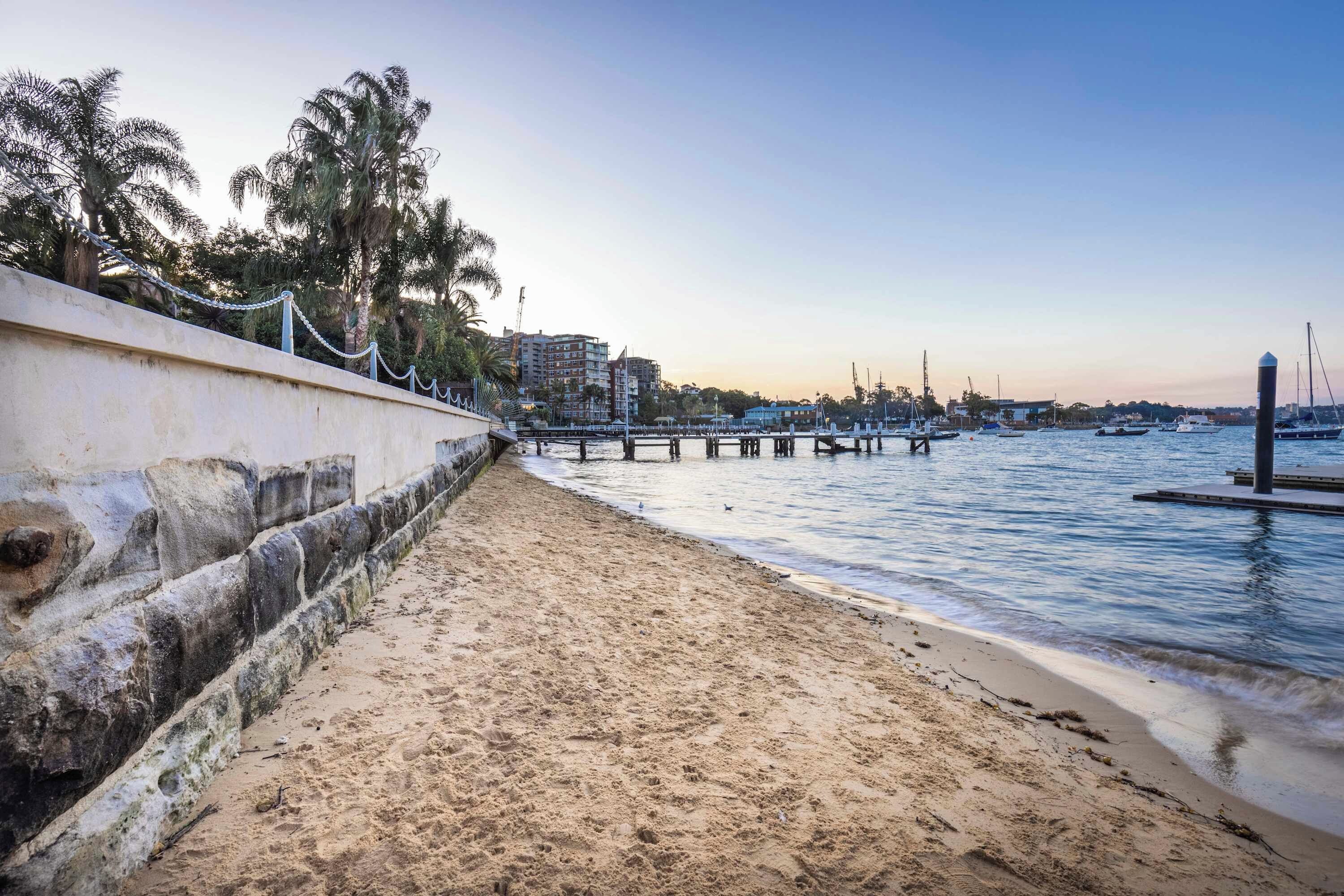 A beach with a concrete walkway, a pier extending into the water, and buildings in the background.