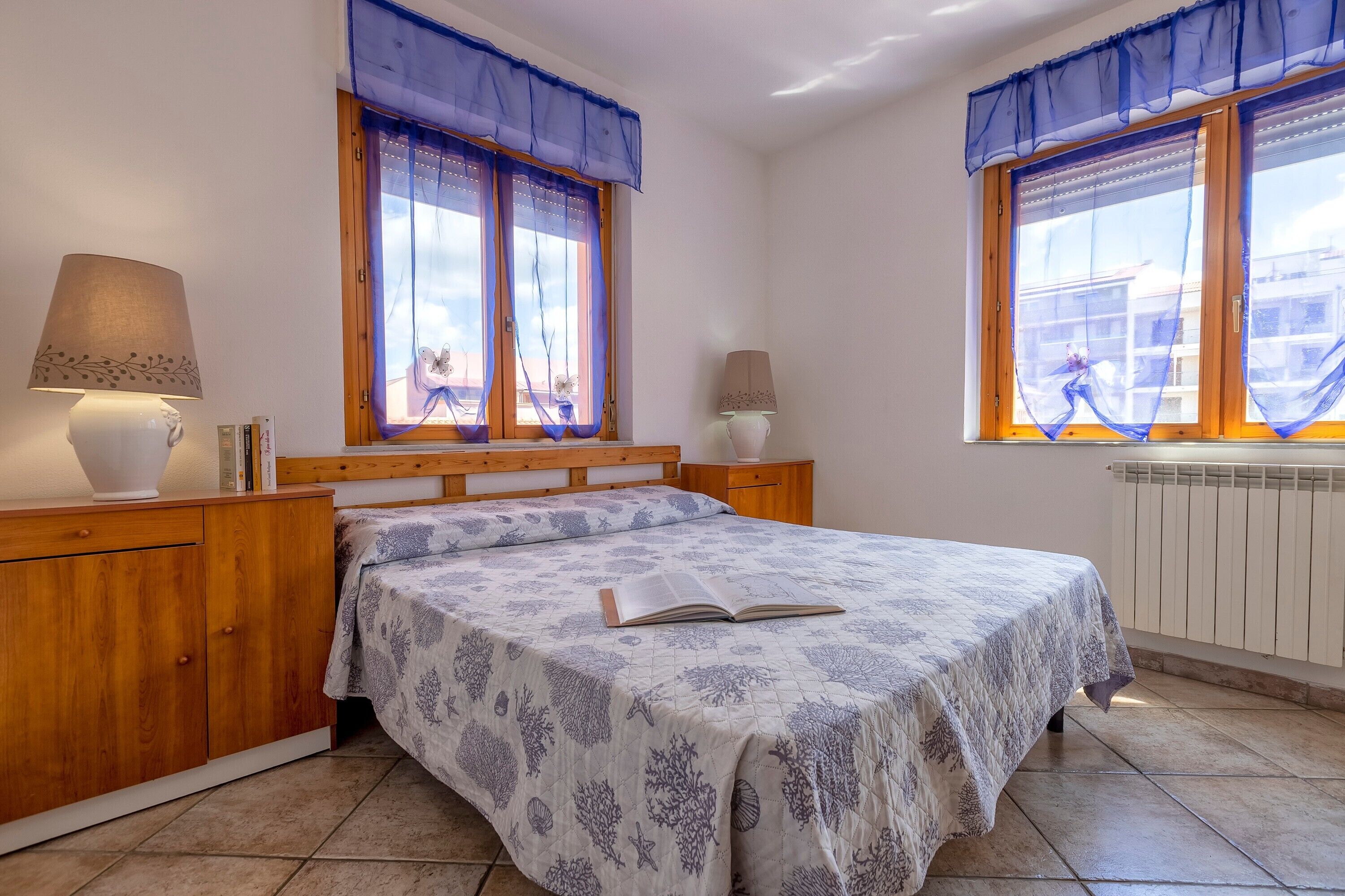 Een slaapkamer met een bed, twee bedlampen, een houten hoofdbord, een nachtkastje, een raam met blauwe gordijnen en een radiator.