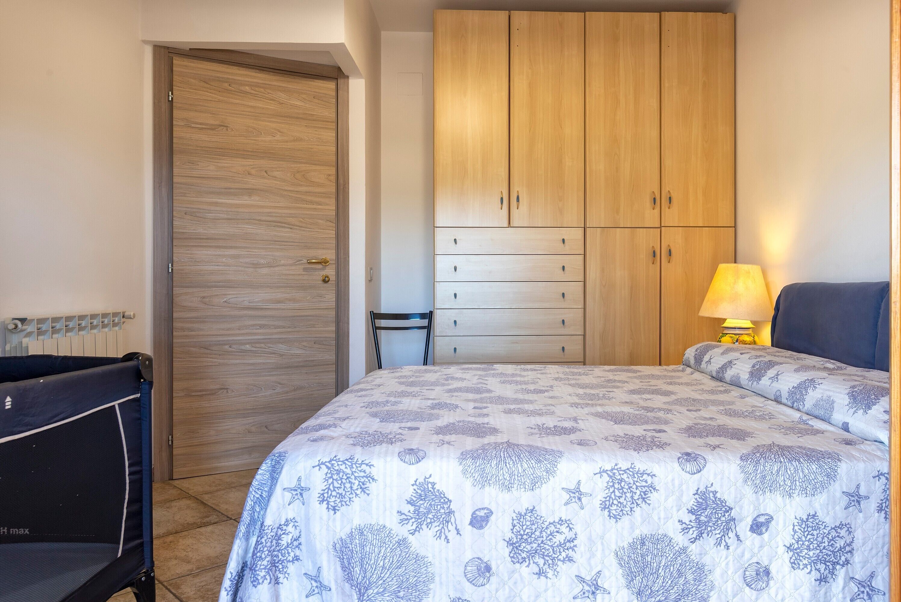 A bedroom with a bed, a wooden wardrobe, a chair, and a bedside table with a lamp.