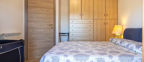 A bedroom with a bed, a wooden wardrobe, a chair, and a bedside table with a lamp.