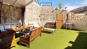 En udendørs terrasse med stenvæg, et træbord med stole, grill og et mindre bord med en stol.