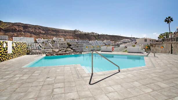 En swimmingpool med flisepyntet solterrasse, en metalstige og udsigt til bygninger samt et bjerg i baggrunden.