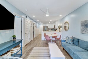 A modern living room with a blue sofa, a dining area with pink chairs, and a kitchen with white cabinets.