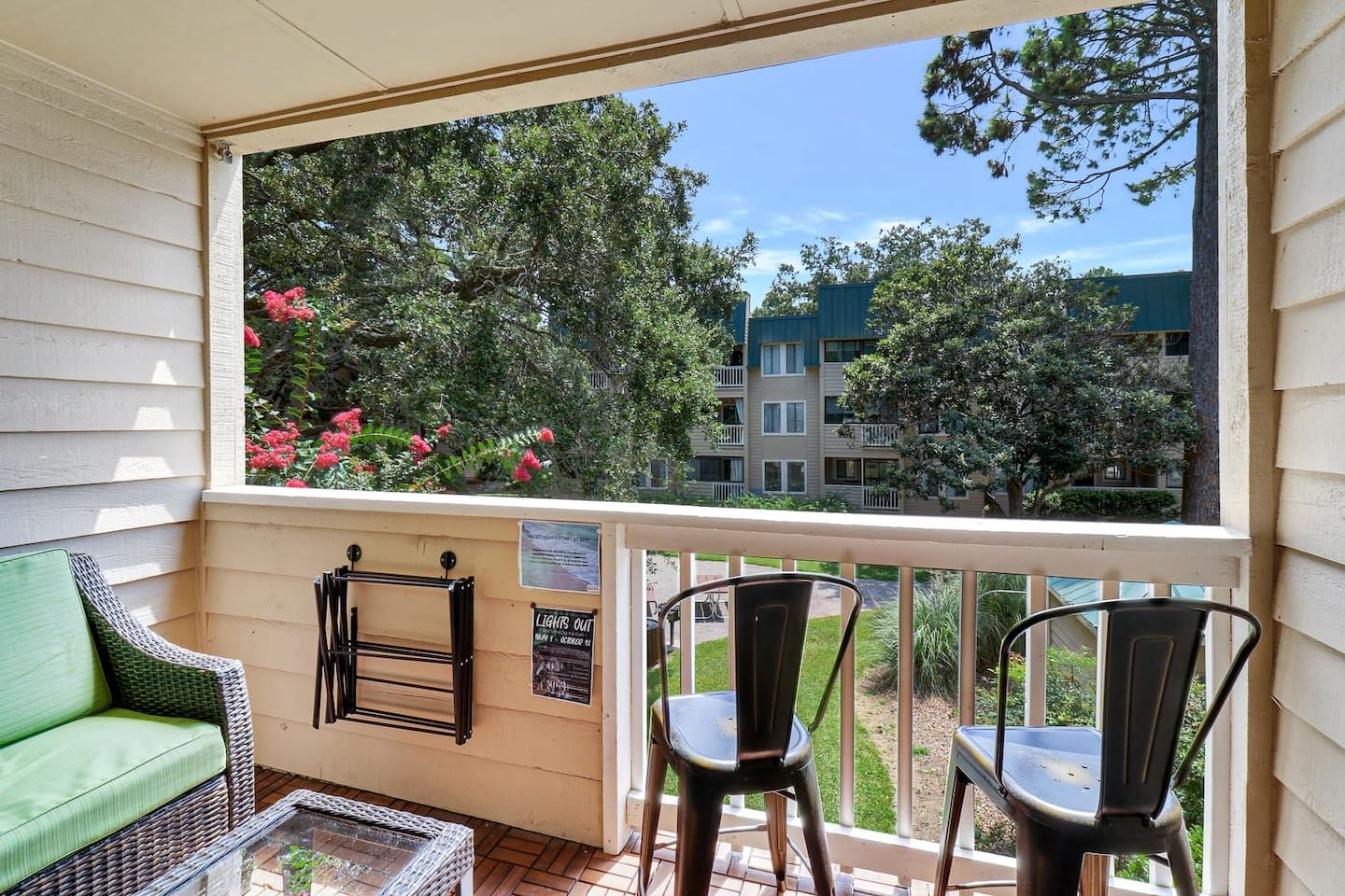 A balcony with wicker furniture, a glass table, and a view of a residential area with trees and buildings.