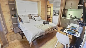 Una camera compatta con un letto, un comodino e una piccola area salotto. Si intravede sullo sfondo un angolo cottura con lavello e tavolo da pranzo.