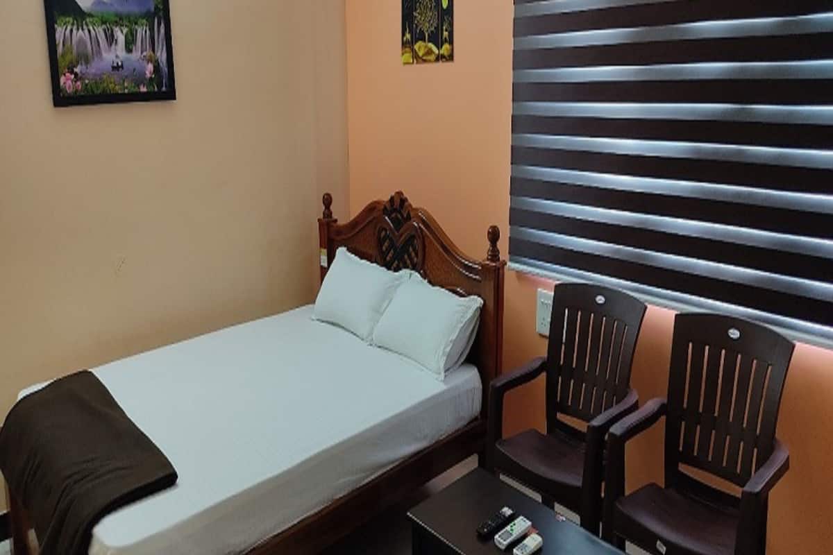 A single bed with a wooden headboard, two chairs, a small table with a remote control, and a window with horizontal blinds.