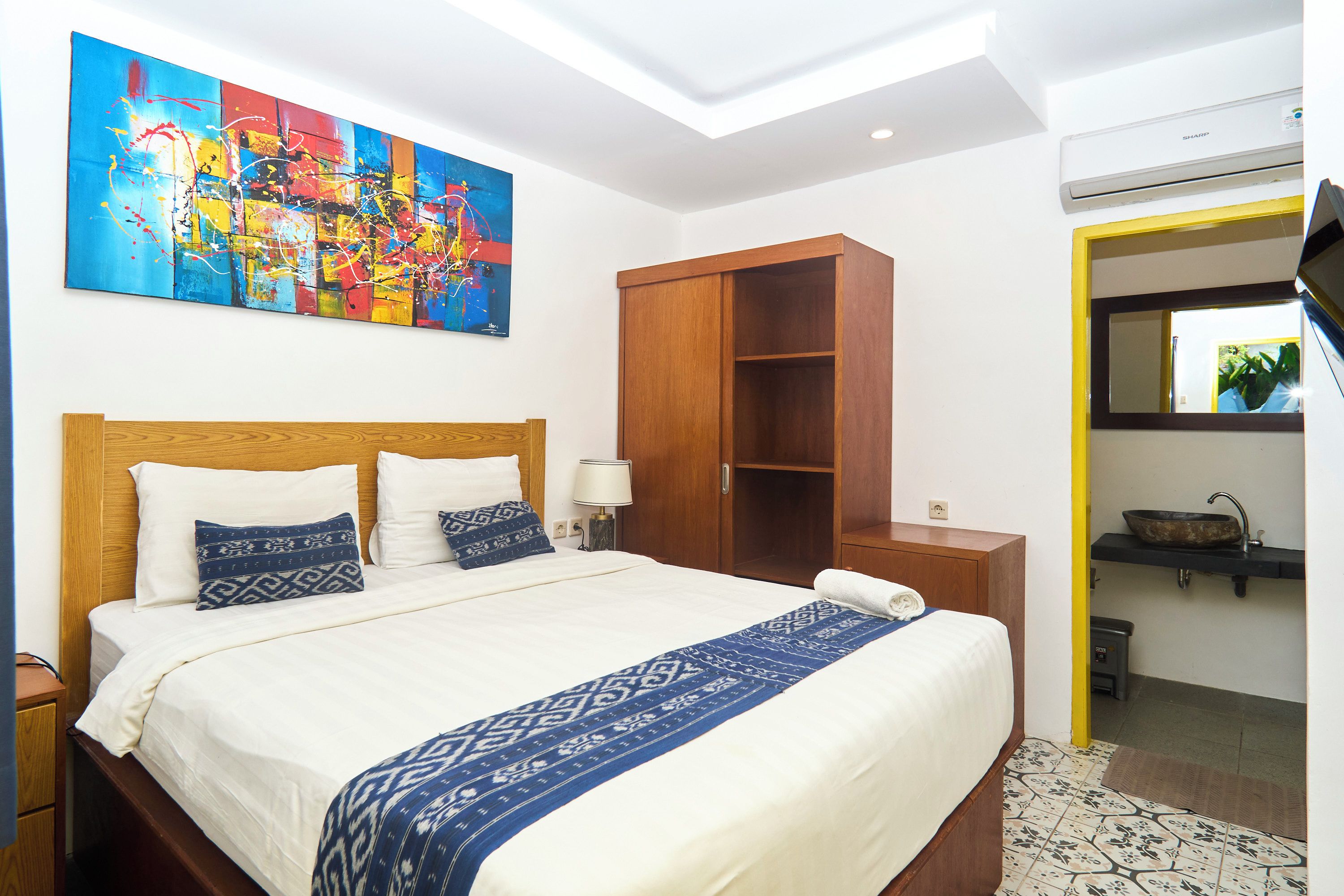 Habitación de hotel con una cama grande, un armario, baño con lavamanos y un cuadro abstracto colorido en la pared.