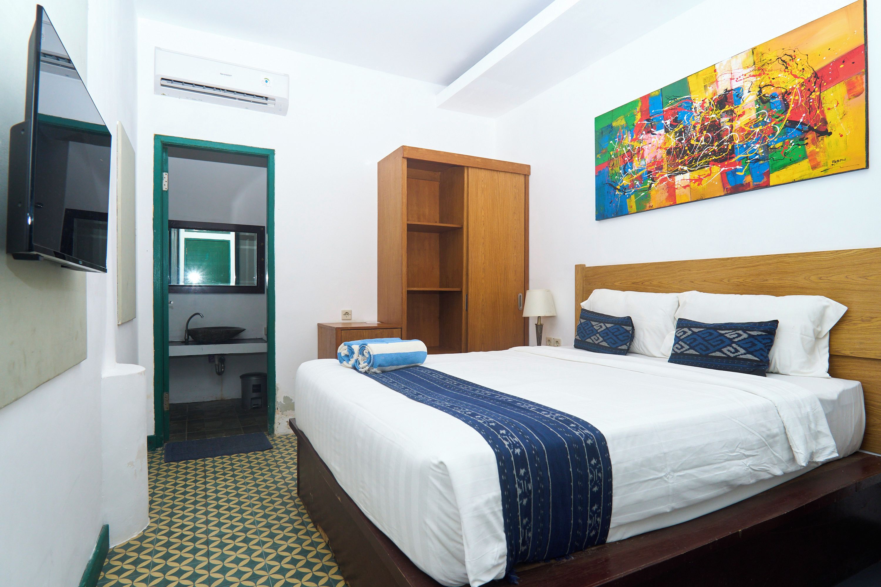 A hotel room with a bed, a television, a painting, a wardrobe, and a bathroom.