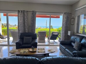 A living room with a view of the sea, a black leather sofa, a wicker chair, and a table with placemats.