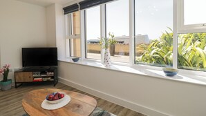 A modern living room with a flat-screen TV, a wooden coffee table with a bowl of fruit, and a large window with a view of greenery. - The Studio (Weston-super-Mare)