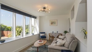 A modern living room with a sofa, a dining table with chairs, a large window with blinds, and a ceiling light fixture. - The Studio (Weston-super-Mare)