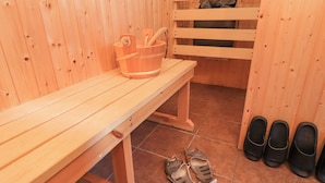 Een houten sauna met banken, een houten emmer en zwarte slippers op de vloer.