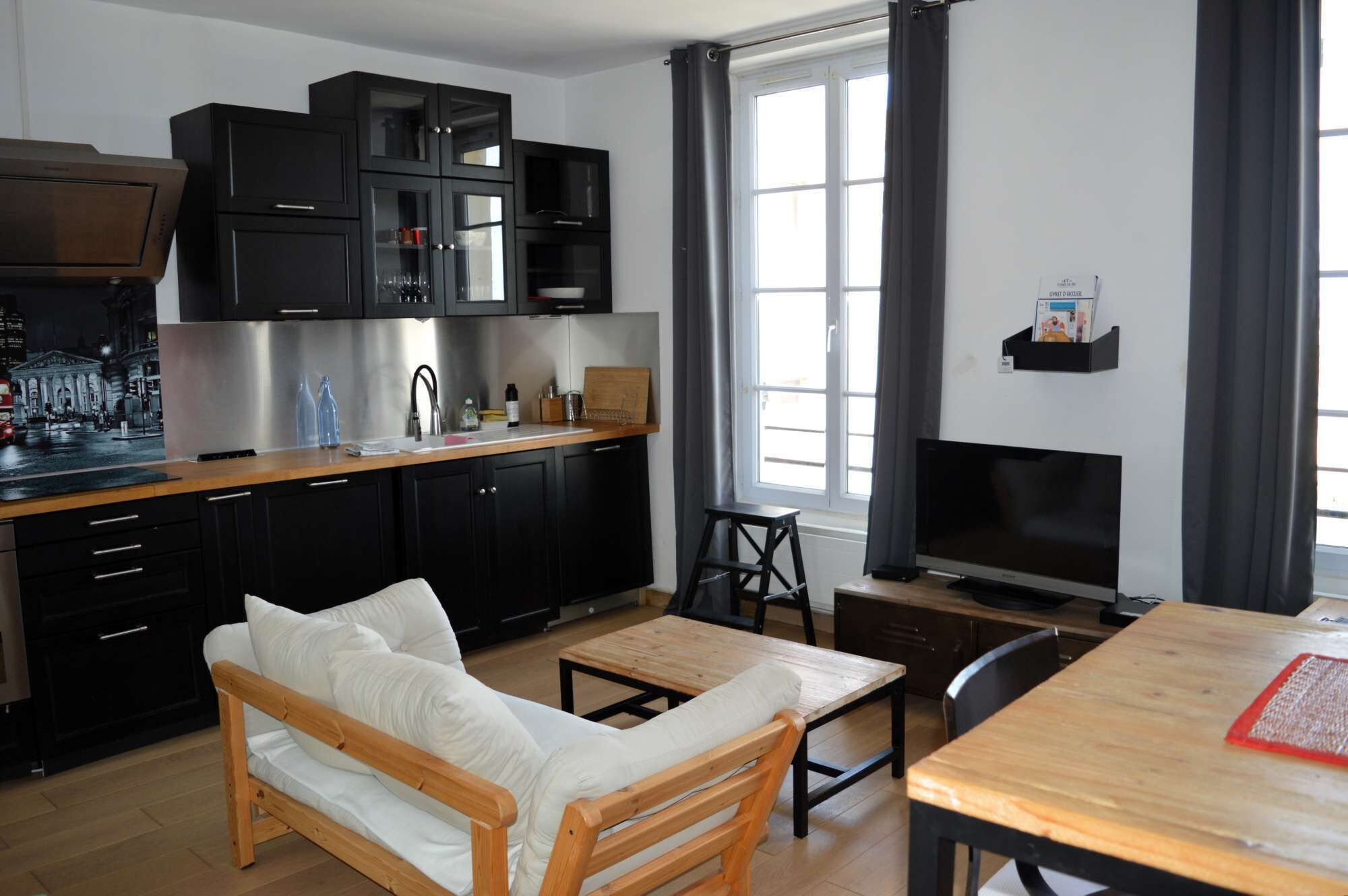 Een moderne keuken met zwarte kasten, een roestvrijstalen koelkast en een houten eettafel met stoelen.