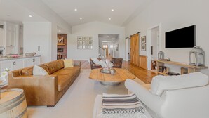 A spacious living area with a large wooden coffee table, leather sofas, and a white armchair. A kitchen with white cabinets and a dining area with a wooden table and chairs are visible in the background.