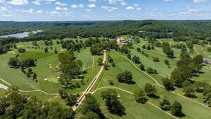 An aerial view of a golf course with multiple fairways and greens, surrounded by dense forests and a lake in the background.