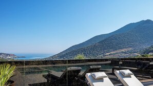 Uma área de piscina no terraço com espreguiçadeiras e vista desobstruída para o mar e montanhas.