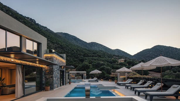 Uma área de piscina moderna ao ar livre, com espreguiçadeiras e guarda-sóis, cercada por montanhas e um edifício com grandes janelas.