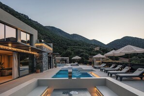 Ein moderner Poolbereich im Freien mit Liegestühlen und Sonnenschirmen, umgeben von Bergen und einem Gebäude mit großen Fenstern.