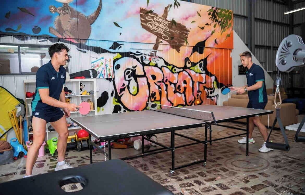 Una mesa de ping pong en una sala con un mural colorido y equipo de ejercicio.