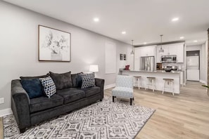 A modern living room with a grey sofa, patterned rug, and a kitchen in the background. - Large 3 bedroom with fenced yard and games! (Columbus)