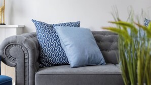 Un divano grigio con cuscini decorati, un quadro appeso al muro e un tavolo rotondo in legno con una pianta in vaso.
