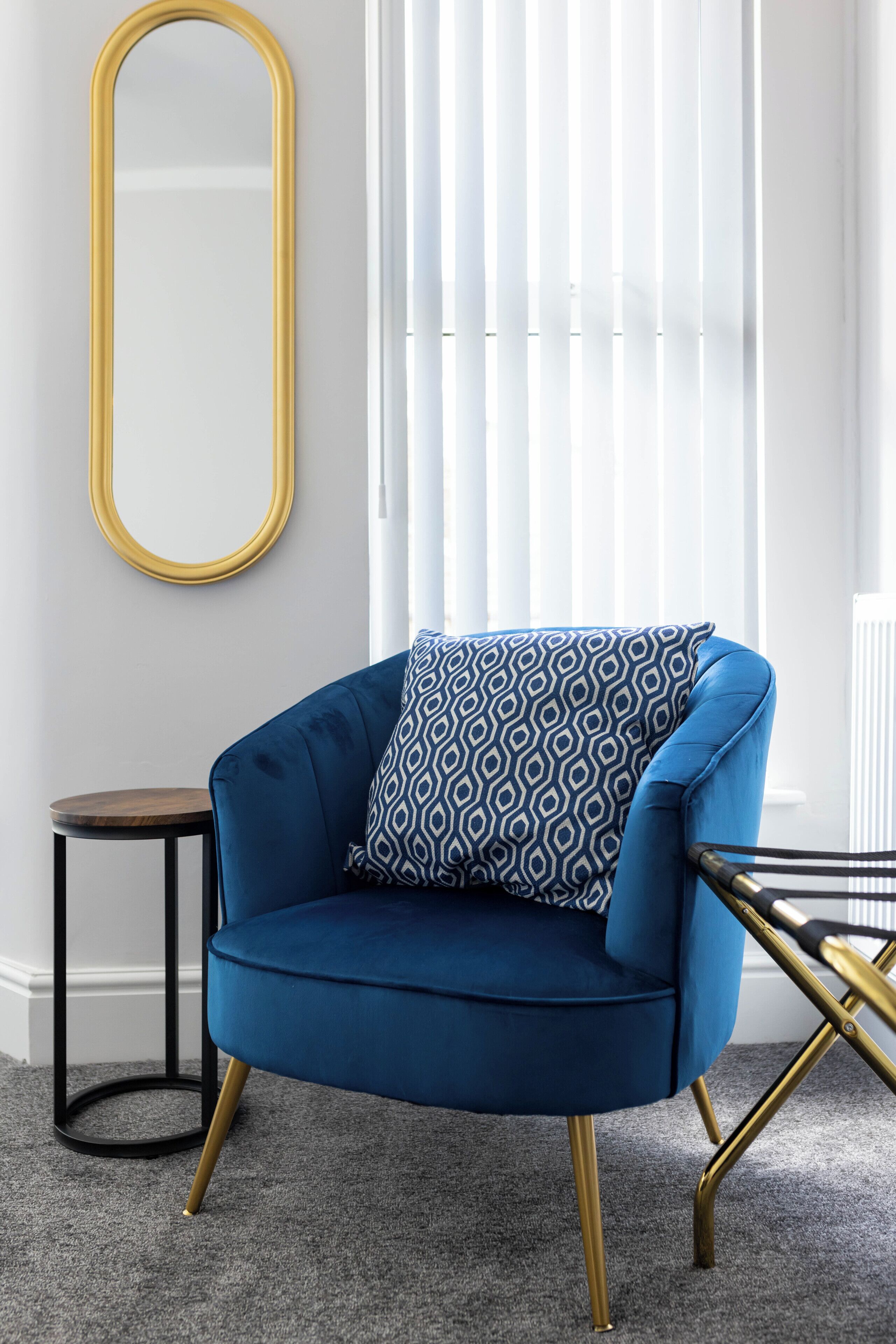 Un sillón azul con un cojín estampado, una mesita redonda pequeña y un espejo con marco dorado sobre una alfombra gris.